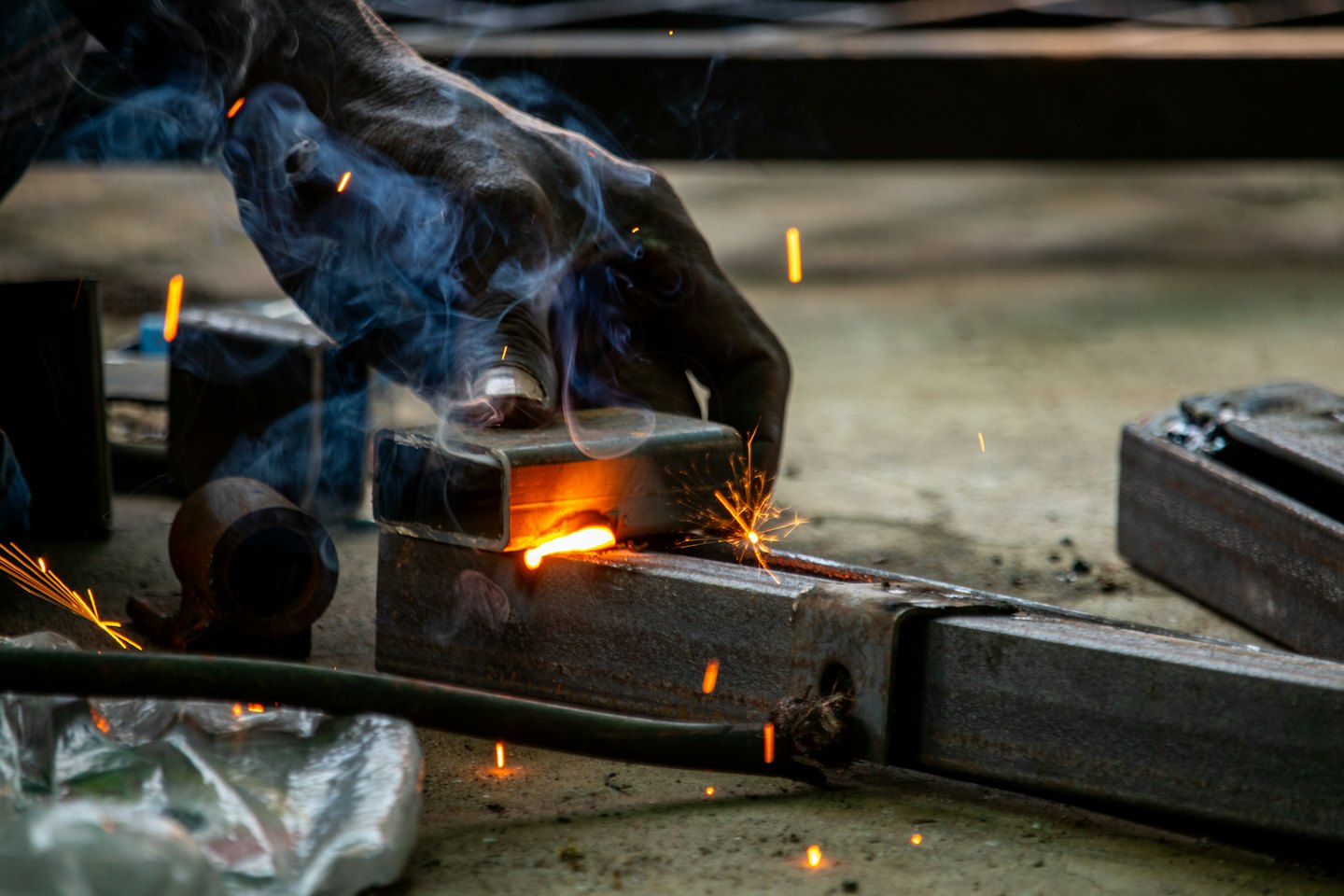 Gloved hand welds metal beams, creating orange sparks and smoke.